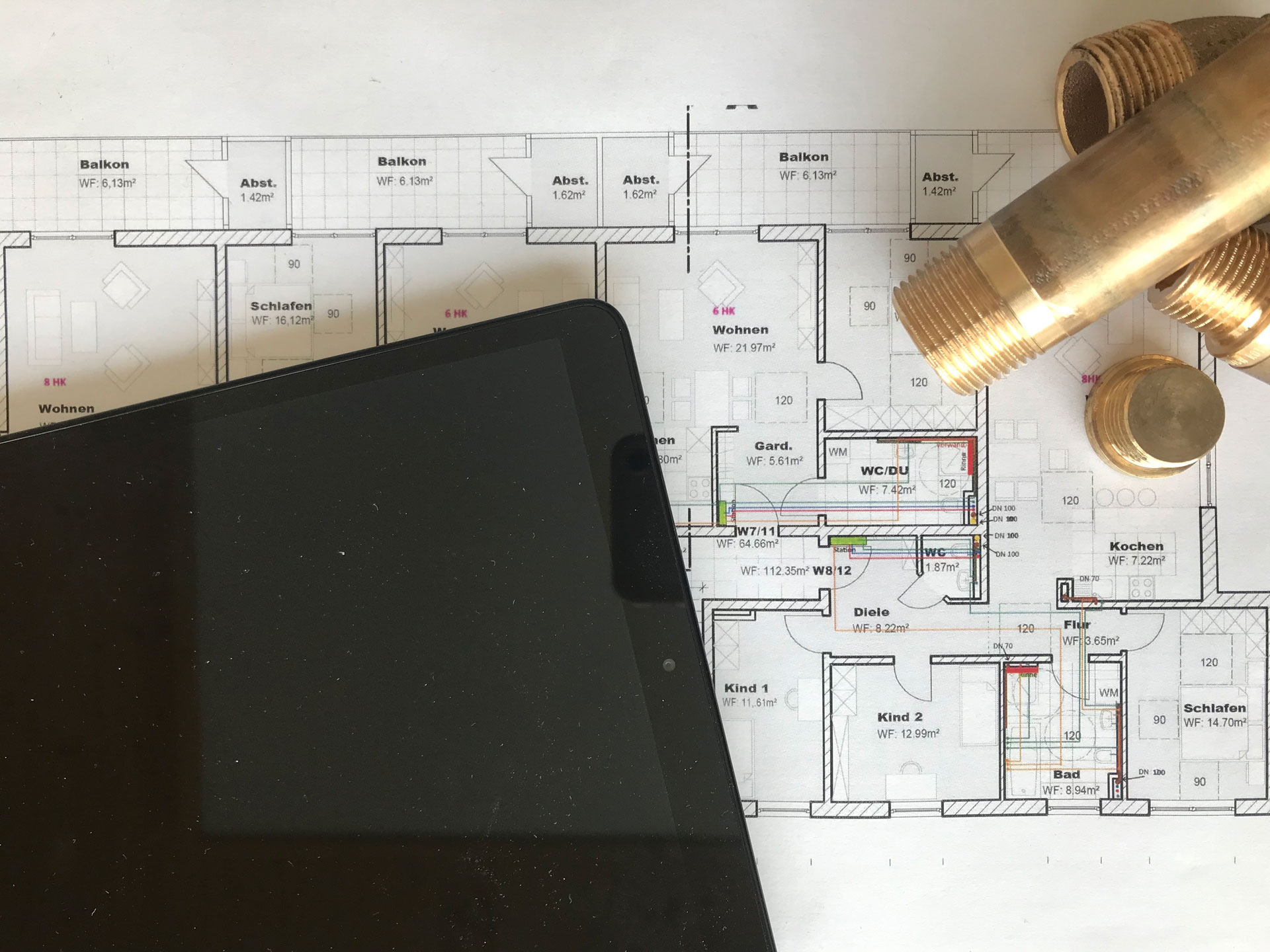 Ein Bauplan einer Wohnung liegt auf einem Tisch, teilweise von einem schwarzen Tablet und mehreren metallischen Rohrverbindungsstücken verdeckt. Der Bauplan zeigt verschiedene Räume, darunter Wohnzimmer, Küche, Schlafzimmer und Badezimmer, sowie Balkone. Die Rohrverbindungsstücke aus Metall und das Tablet symbolisieren eine technische Planung, möglicherweise im Zusammenhang mit der Installation von Sanitär- oder Heizsystemen.